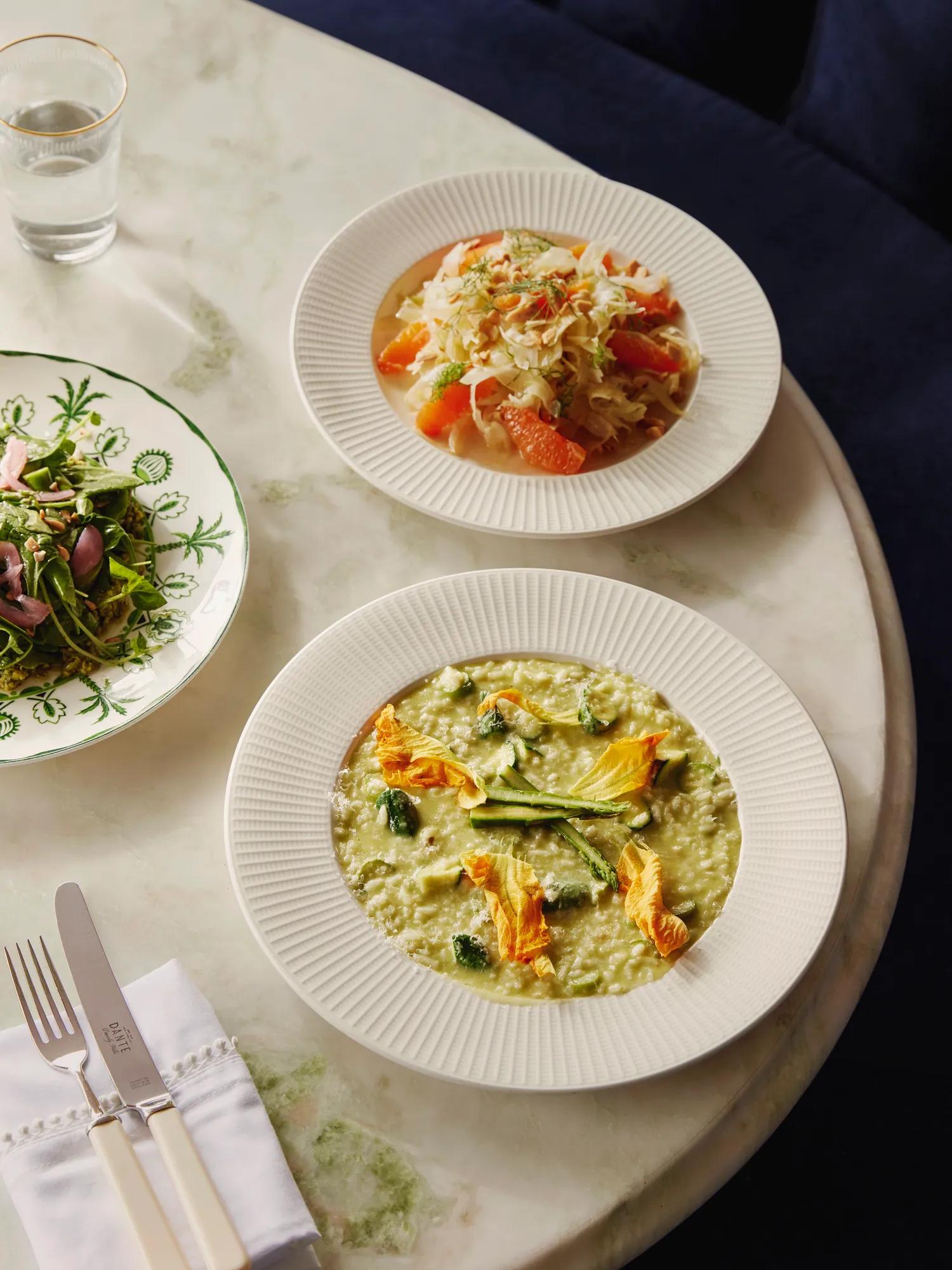 Marble table with three dishes: citrus-fennel salad, green risotto with zucchini blossoms, and a leafy green salad.