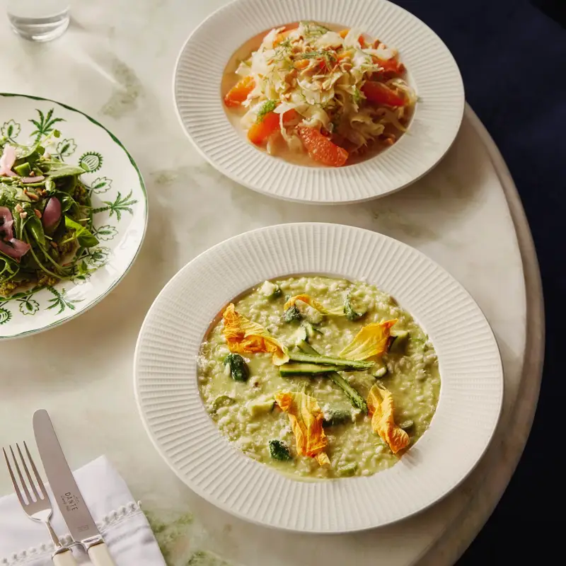 Marble table with three dishes: citrus-fennel salad, green risotto with zucchini blossoms, and a leafy green salad.