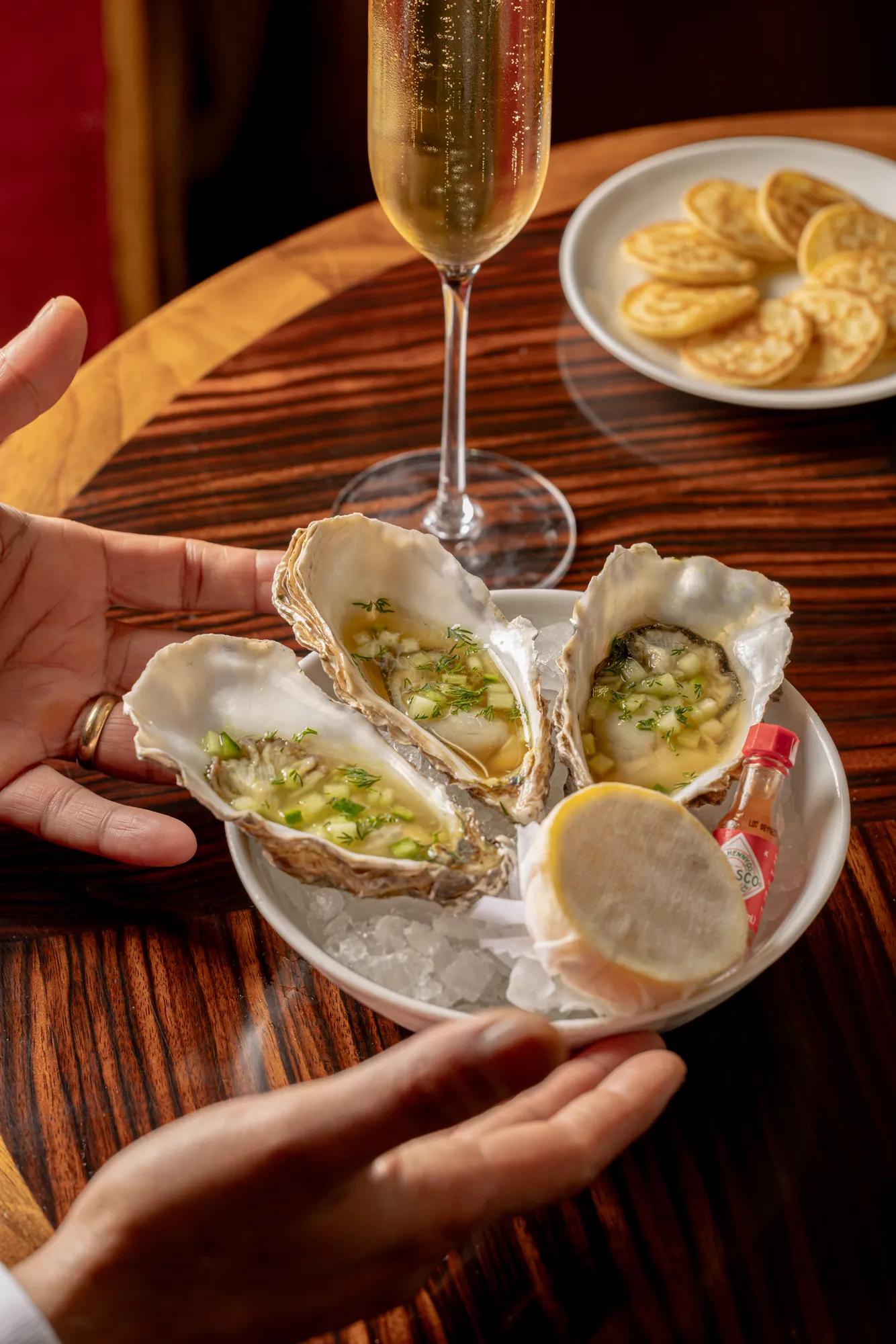 Three oysters on ice with garnish, lemon, and hot sauce, held by hands on wooden table with champagne flute and crackers nearby.