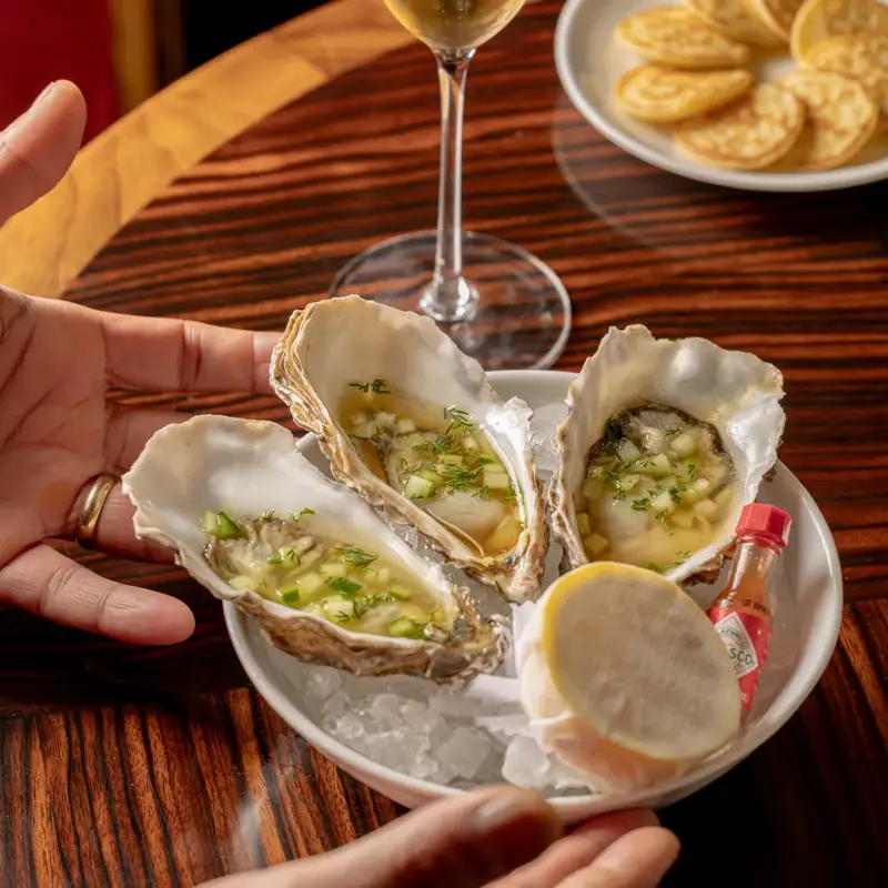 Three oysters on ice with garnish, lemon, and hot sauce, held by hands on wooden table with champagne flute and crackers nearby.