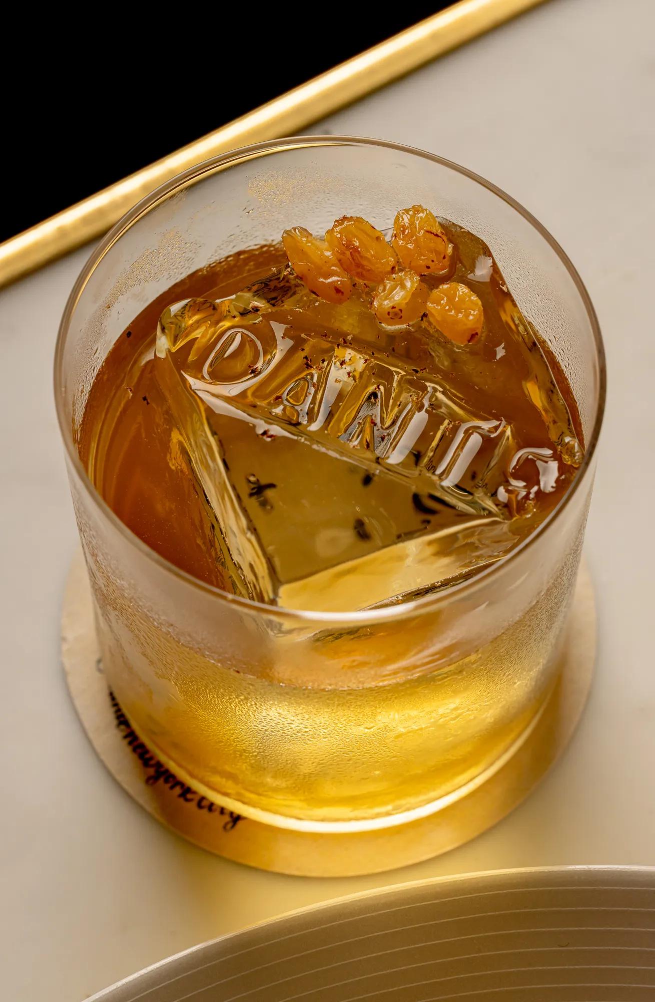 Glass of amber-coloured cocktail with a large ice cube and citrus garnish, served on a coaster.
