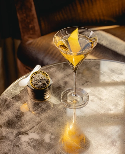 A martini with a lemon twist on a reflective table, served beside a small tin of caviar and a white spoon.
