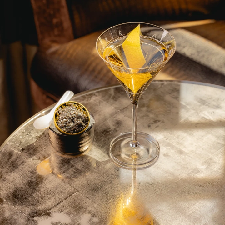 A martini with a lemon twist on a reflective table, served beside a small tin of caviar and a white spoon.