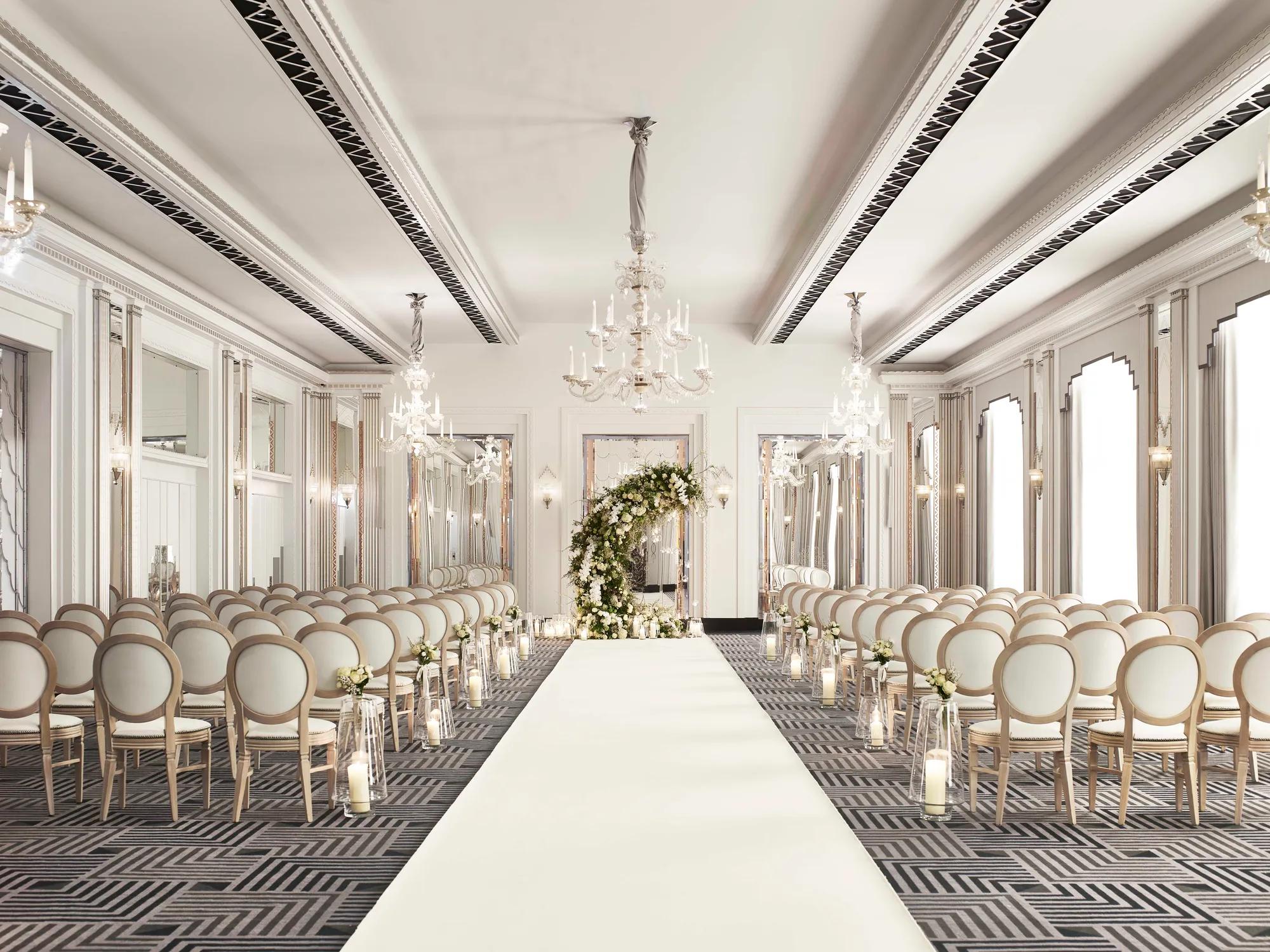Elegant wedding ceremony room with chandelier lighting, white aisle runner, floral arch, and rows of cream chairs.
