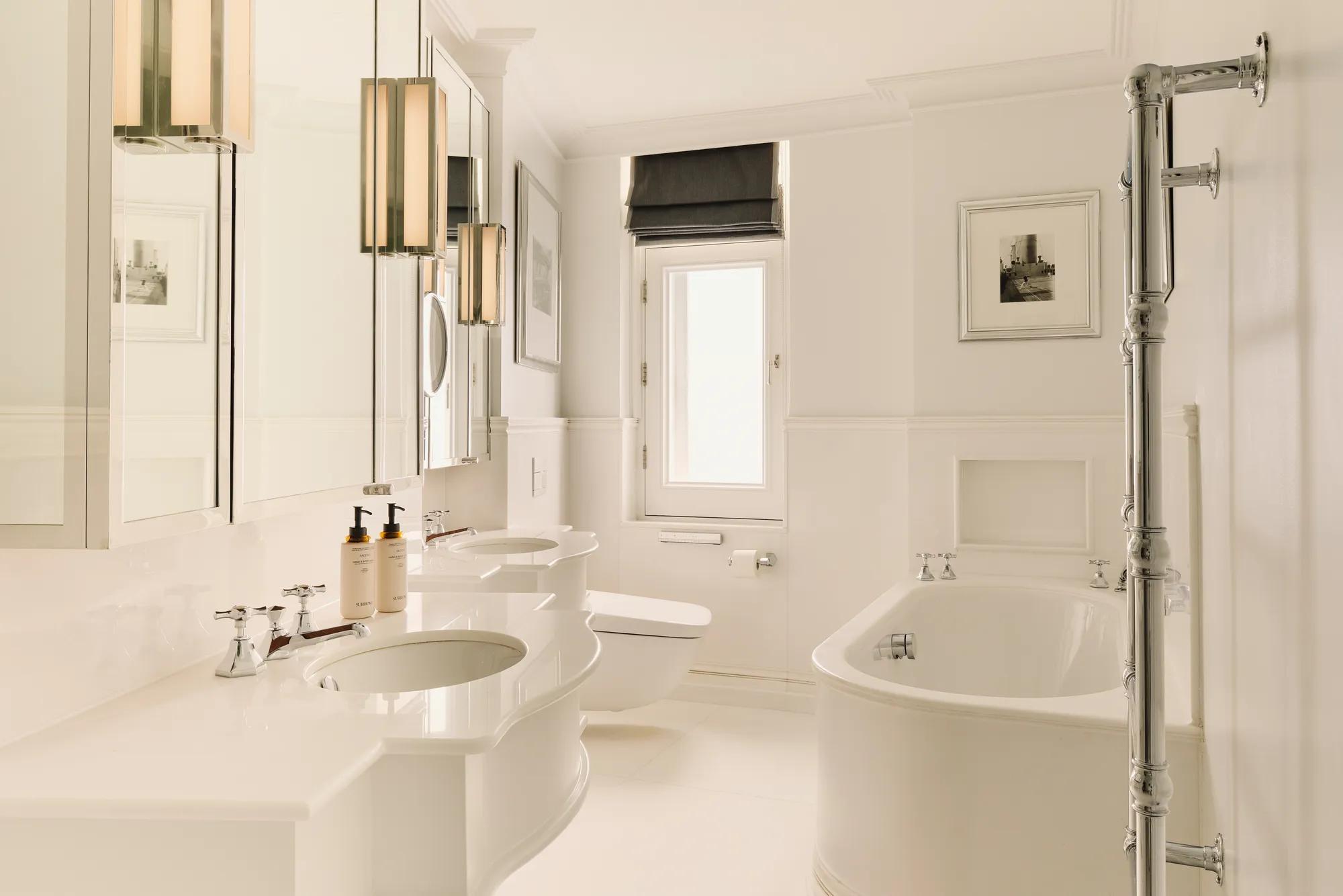 Bright white bathroom with double sinks, rounded bathtub, chrome towel rail, and a small window with a dark blind.