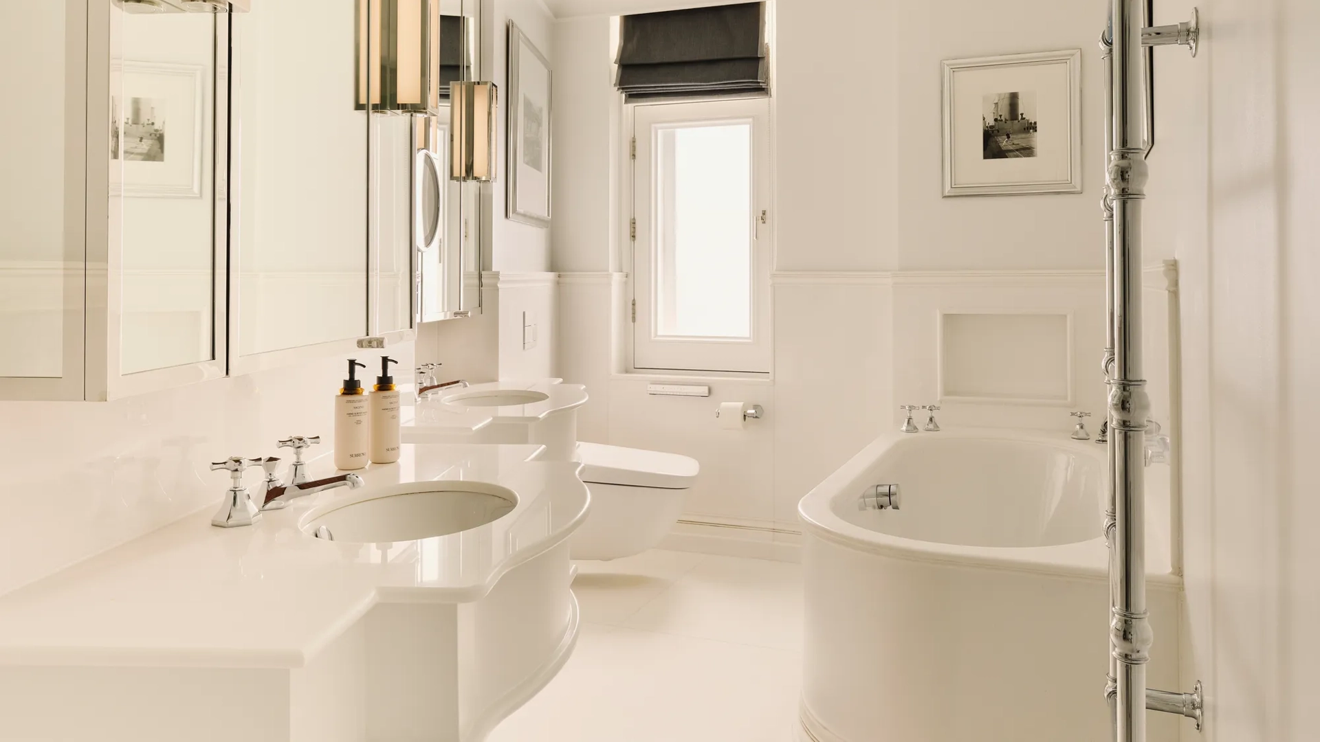 Bright white bathroom with double sinks, rounded bathtub, chrome towel rail, and a small window with a dark blind.
