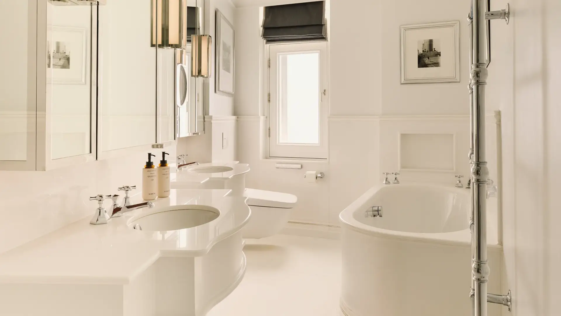 Bright white bathroom with double sinks, rounded bathtub, chrome towel rail, and a small window with a dark blind.