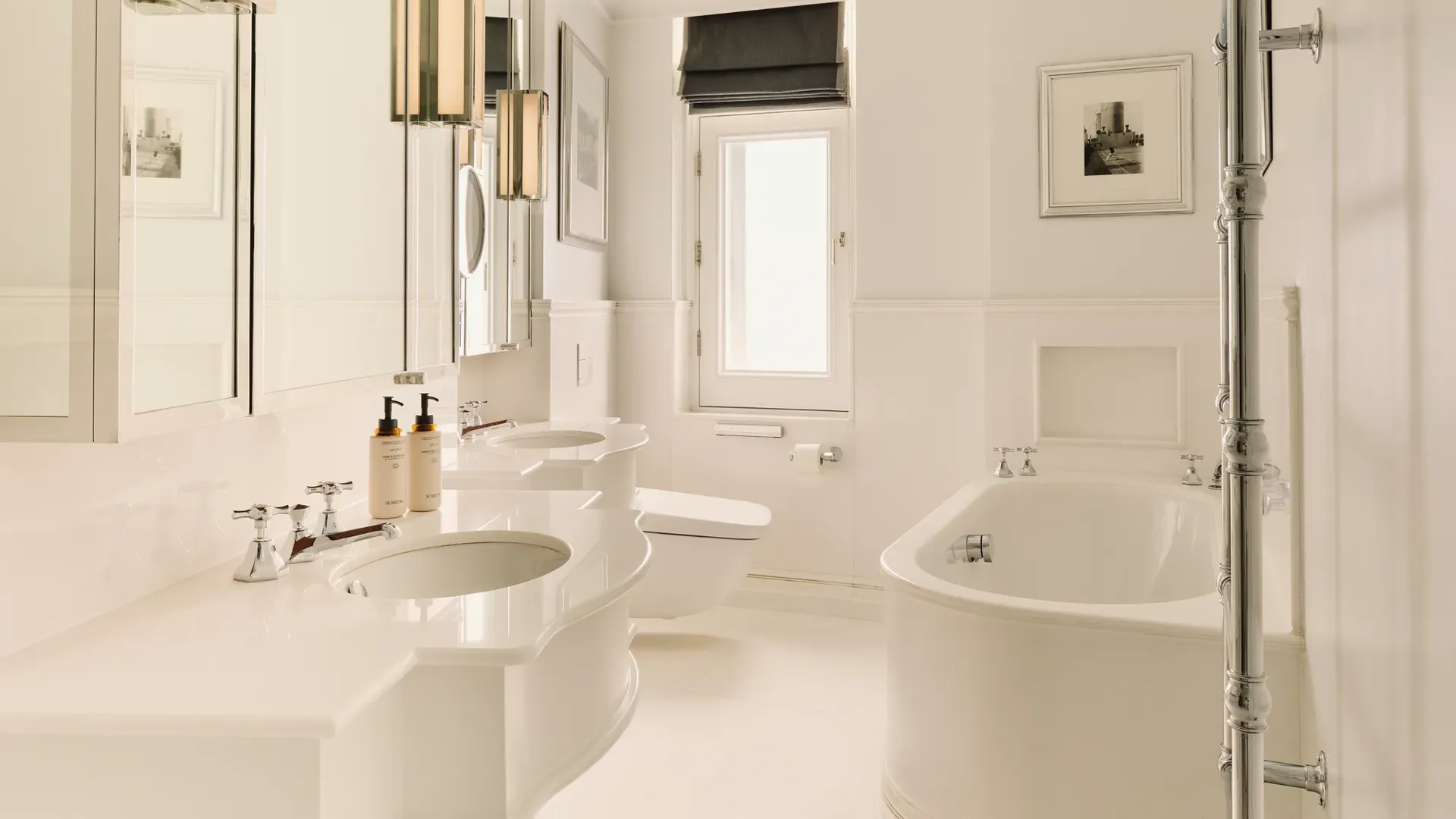 Bright white bathroom with double sinks, rounded bathtub, chrome towel rail, and a small window with a dark blind.