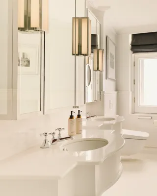 Bright white bathroom with double sinks, rounded bathtub, chrome towel rail, and a small window with a dark blind.