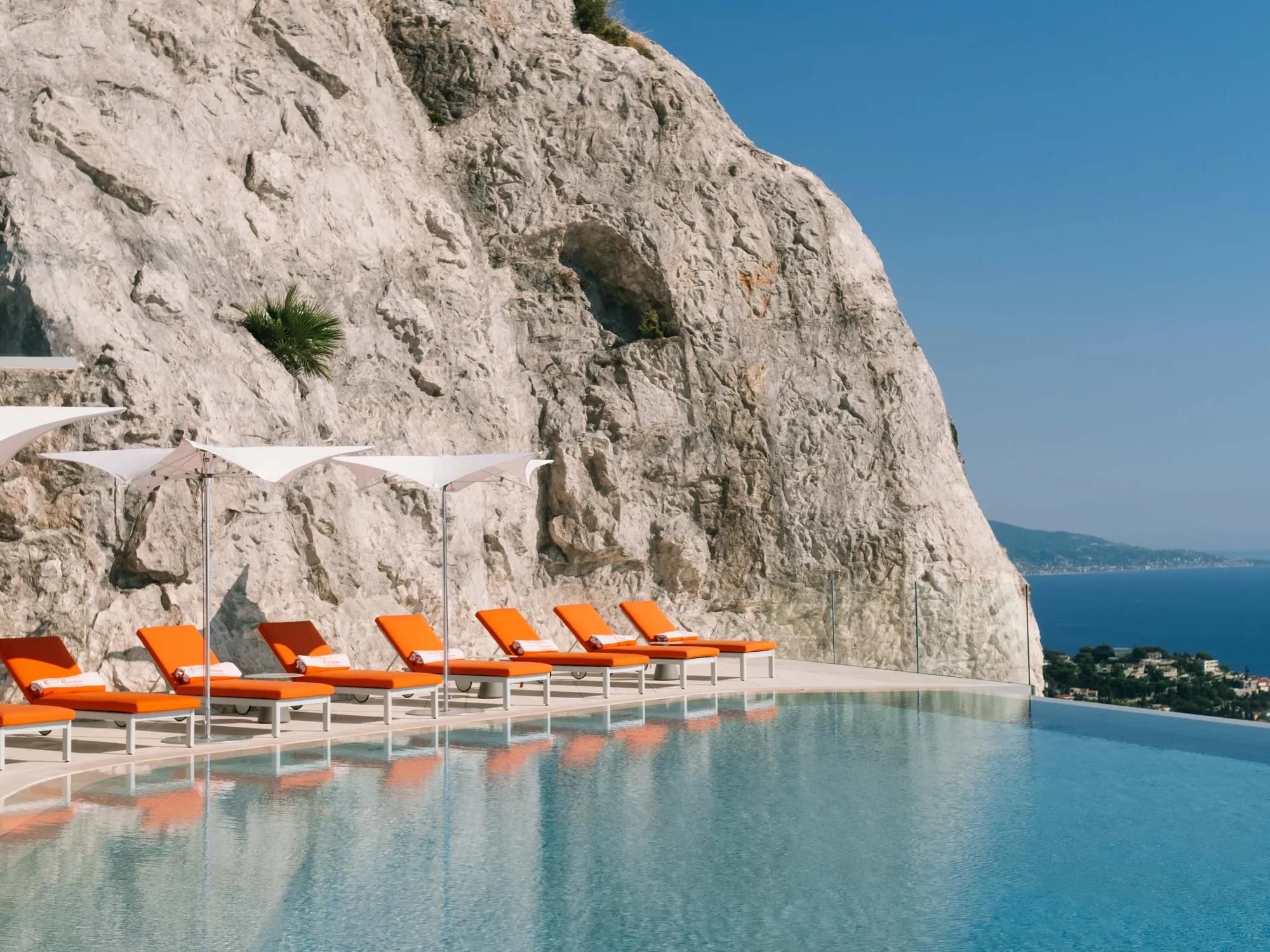 Piscine à débordement adossée à une falaise spectaculaire avec des transats orange vif et des parasols, surplombant la côte méditerranéenne.