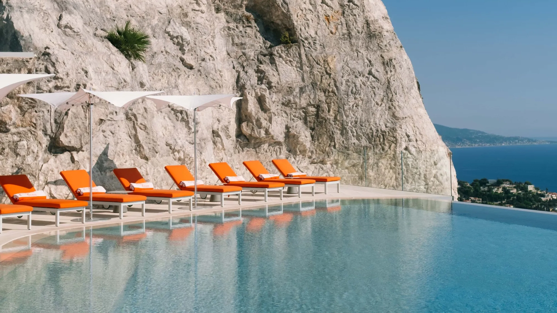 Infinity pool set against a dramatic cliff face with bright orange sun loungers and parasols, overlooking the Mediterranean coastline.