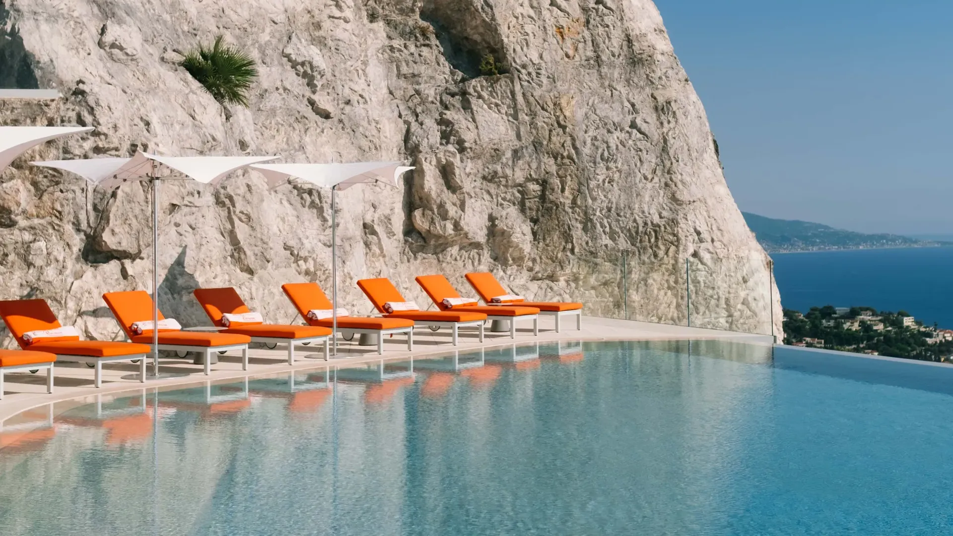Piscine à débordement adossée à une falaise spectaculaire avec des transats orange vif et des parasols, surplombant la côte méditerranéenne.