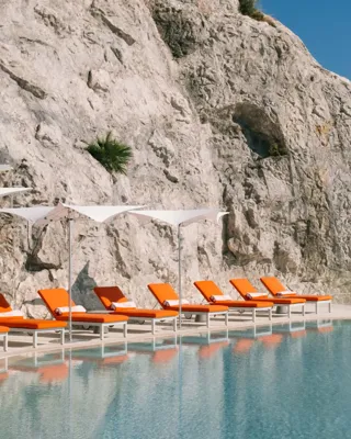 Piscine à débordement adossée à une falaise spectaculaire avec des transats orange vif et des parasols, surplombant la côte méditerranéenne.