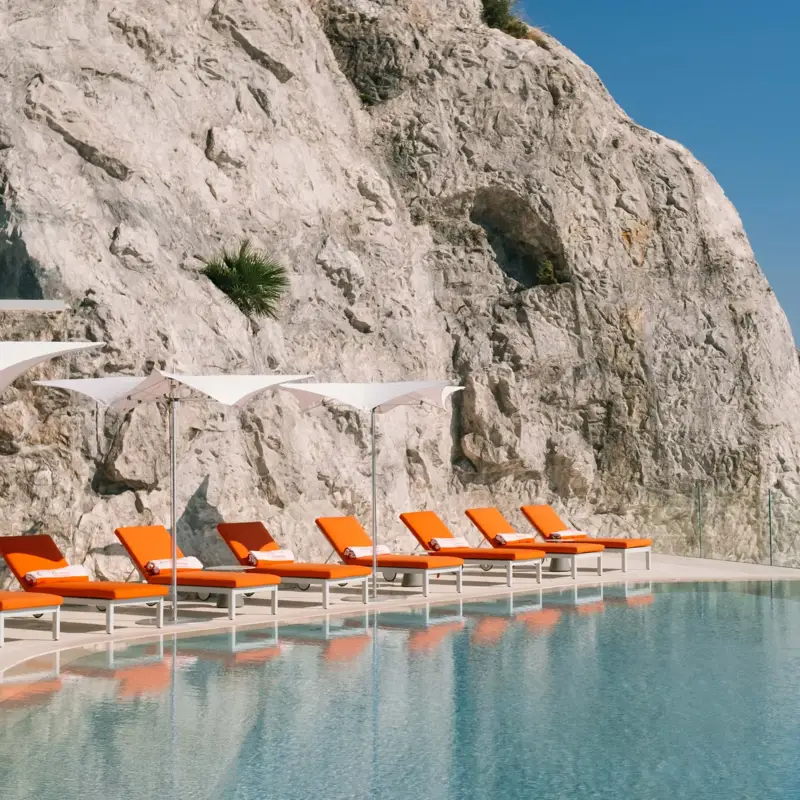 Infinity pool set against a dramatic cliff face with bright orange sun loungers and parasols, overlooking the Mediterranean coastline.