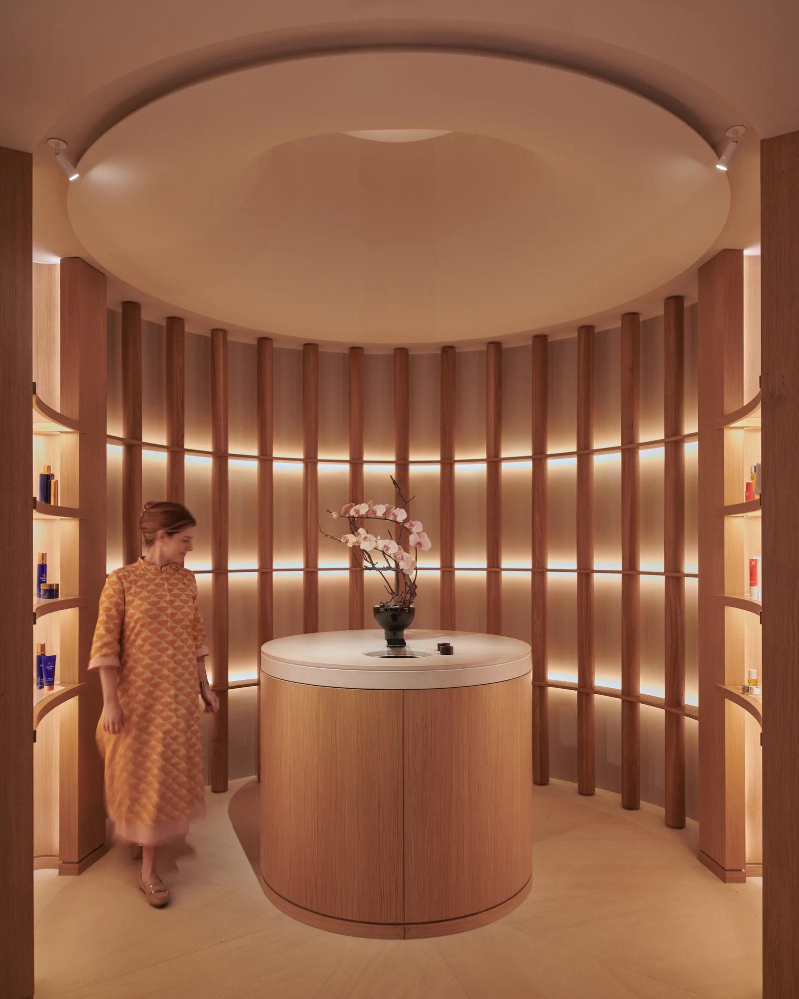 A woman in a patterned dress stands in a softly lit circular alcove at The Maybourne Riviera, featuring curved wooden panels, warm lighting, and a round table with orchids.