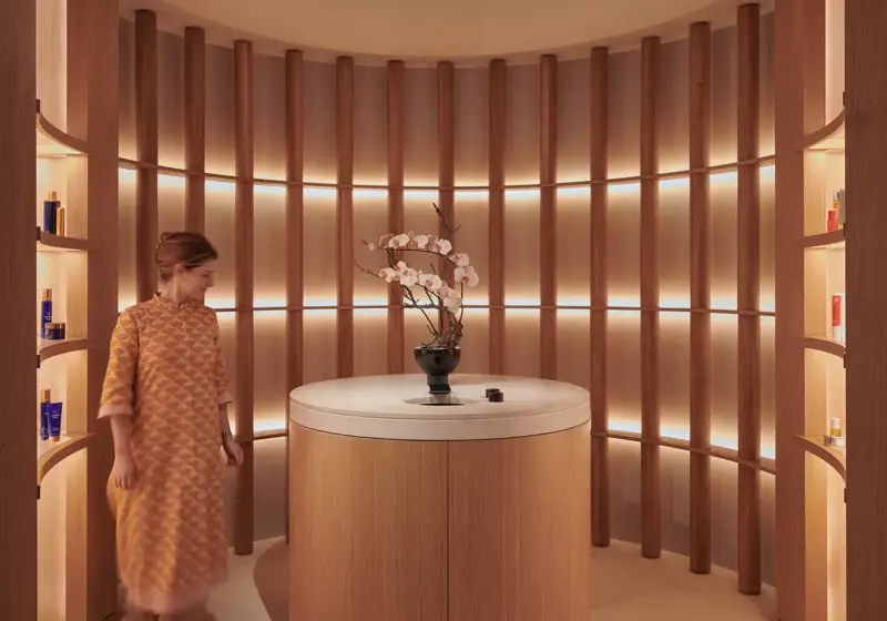 A woman in a patterned dress stands in a softly lit circular alcove at The Maybourne Riviera, featuring curved wooden panels, warm lighting, and a round table with orchids.
