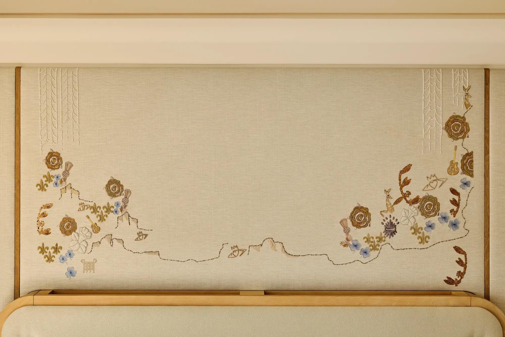 Close-up of a decorative embroidered wall panel featuring delicate floral motifs in gold, blue, and cream tones above a light wood headboard.