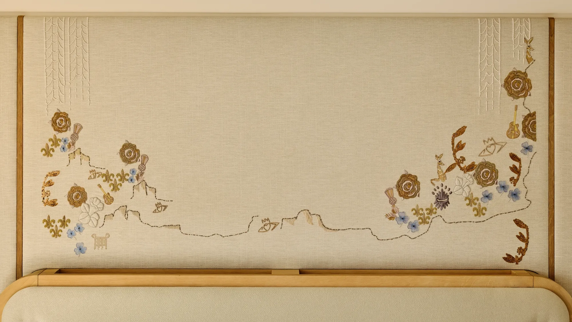 Close-up of a decorative embroidered wall panel featuring delicate floral motifs in gold, blue, and cream tones above a light wood headboard.