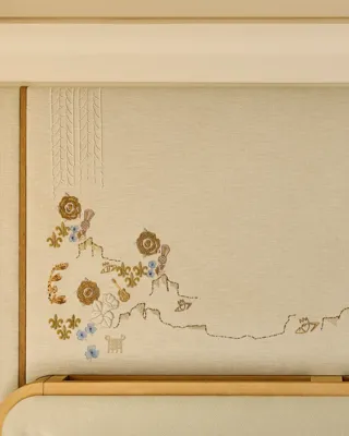 Close-up of a decorative embroidered wall panel featuring delicate floral motifs in gold, blue, and cream tones above a light wood headboard.