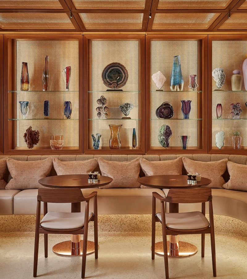 Lounge seating area with two round tables and chairs, backed by illuminated shelves displaying decorative glass art.
