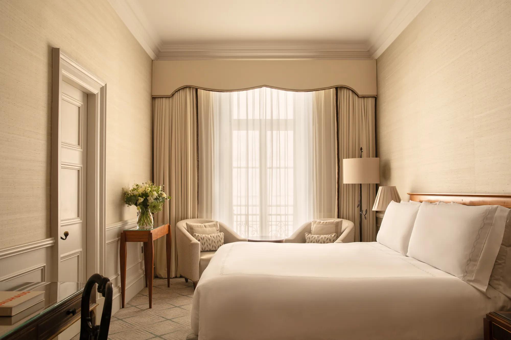Bright guest bedroom with a king bed, two armchairs by a tall window, a writing desk and fresh flowers in soft neutral tones.