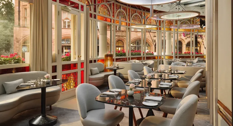 Elegant restaurant with curved banquettes, marble tables, and stained glass windows overlooking a historic street.