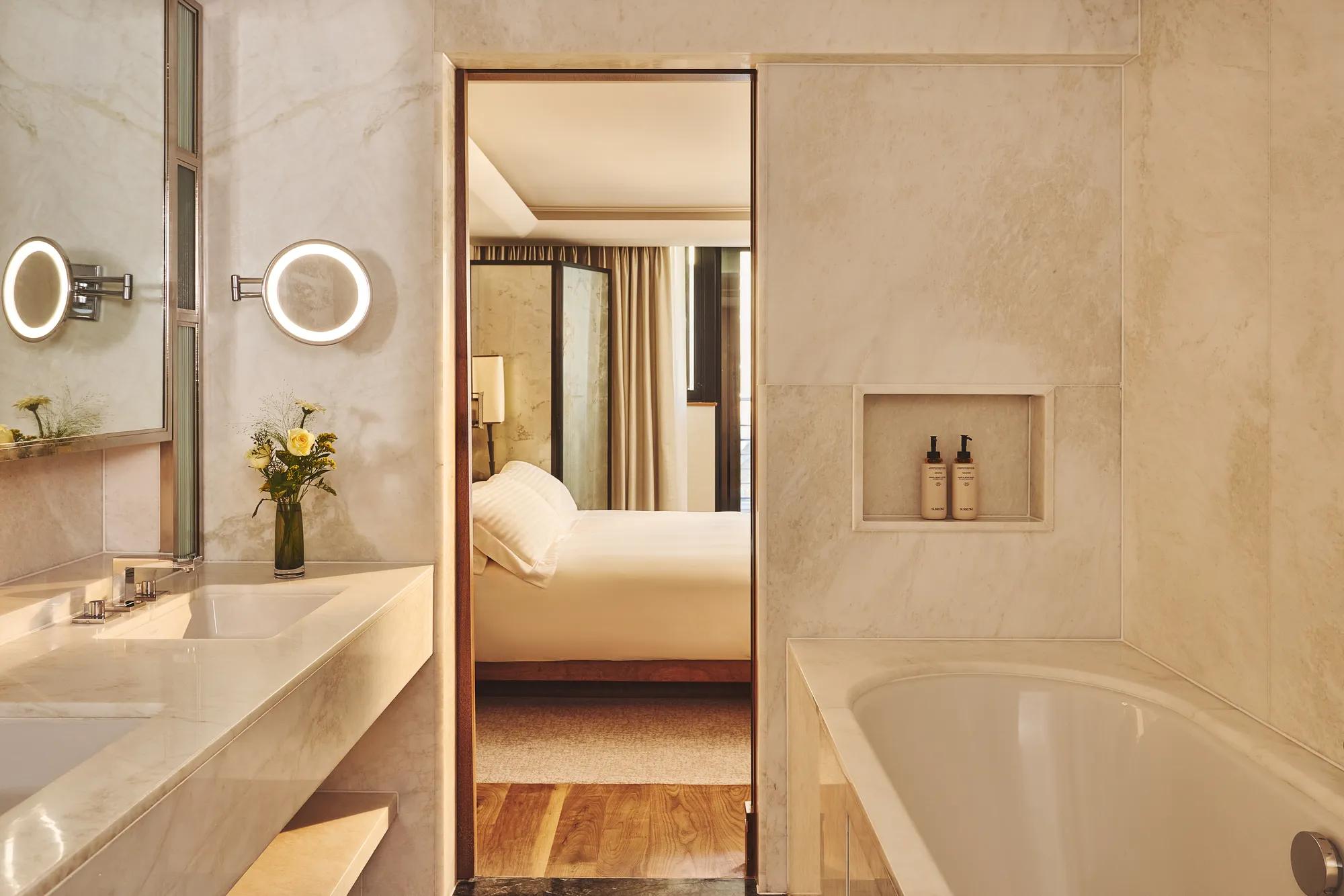 Marble bathroom with a large vanity and tub, looking through an open doorway to a softly lit bedroom with a neatly made bed.