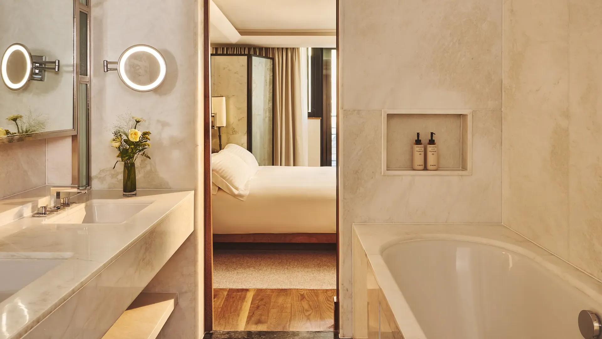 Marble bathroom with a large vanity and tub, looking through an open doorway to a softly lit bedroom with a neatly made bed.