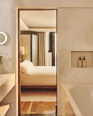 Marble bathroom with a large vanity and tub, looking through an open doorway to a softly lit bedroom with a neatly made bed.