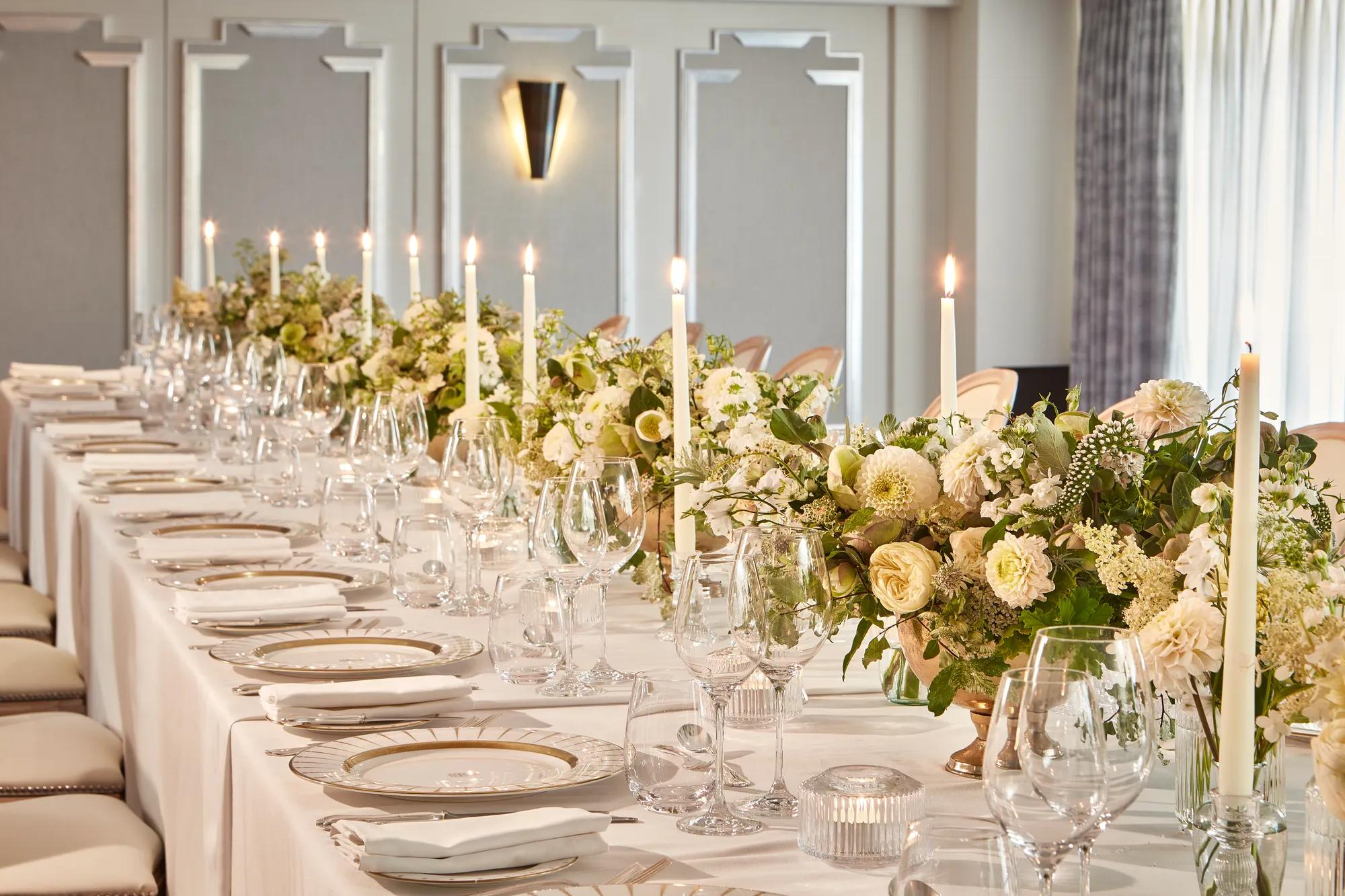 Elegant banquet table set with white linens, gold-rim plates, glassware, candles, and cream floral centrepieces in a bright room
