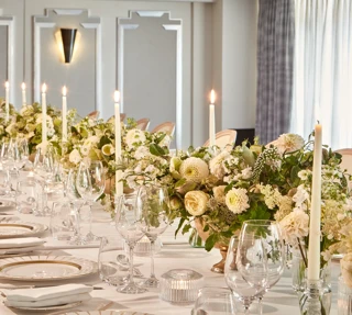 Elegant banquet table set with white linens, gold-rim plates, glassware, candles, and cream floral centrepieces in a bright room