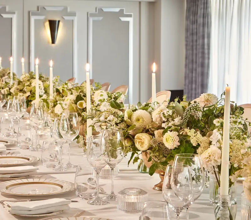 Elegant banquet table set with white linens, gold-rim plates, glassware, candles, and cream floral centrepieces in a bright room