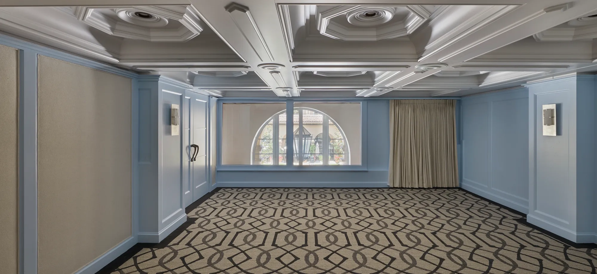 Empty Wilshire Room with pale blue paneling, coffered ceiling, arched window, and geometric patterned carpet.