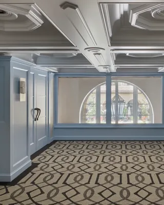 Empty Wilshire Room with pale blue paneling, coffered ceiling, arched window, and geometric patterned carpet.