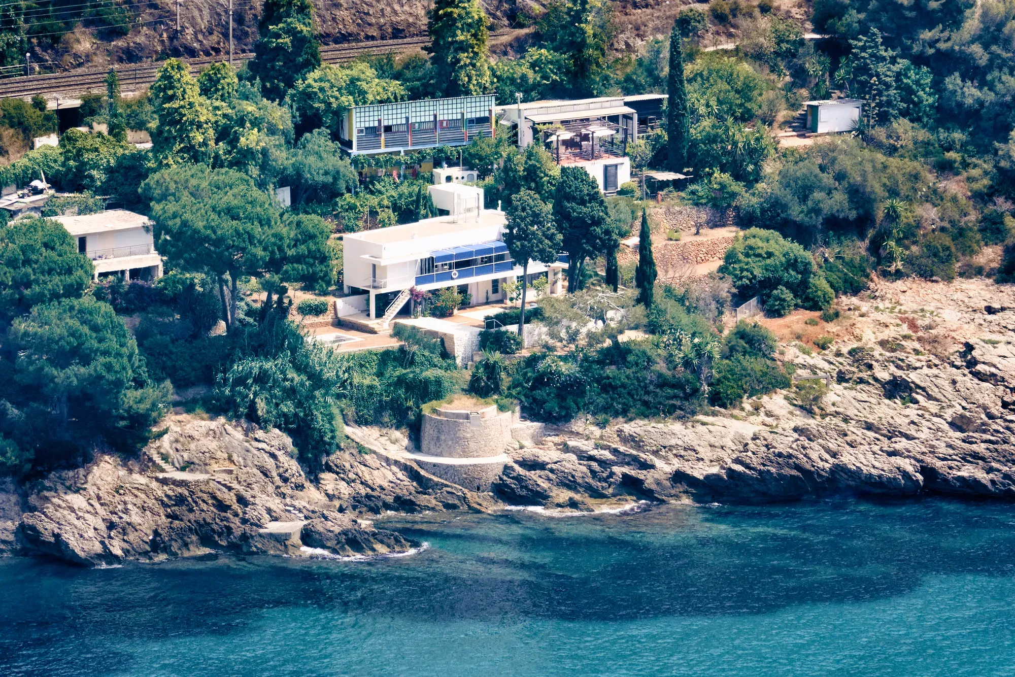 A modern villa perched above clear blue water, with terraces overlooking the sea and sunlight reflecting off the waves below