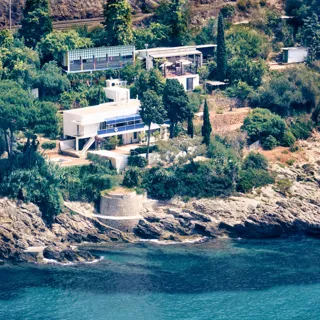 A modern villa perched above clear blue water, with terraces overlooking the sea and sunlight reflecting off the waves below