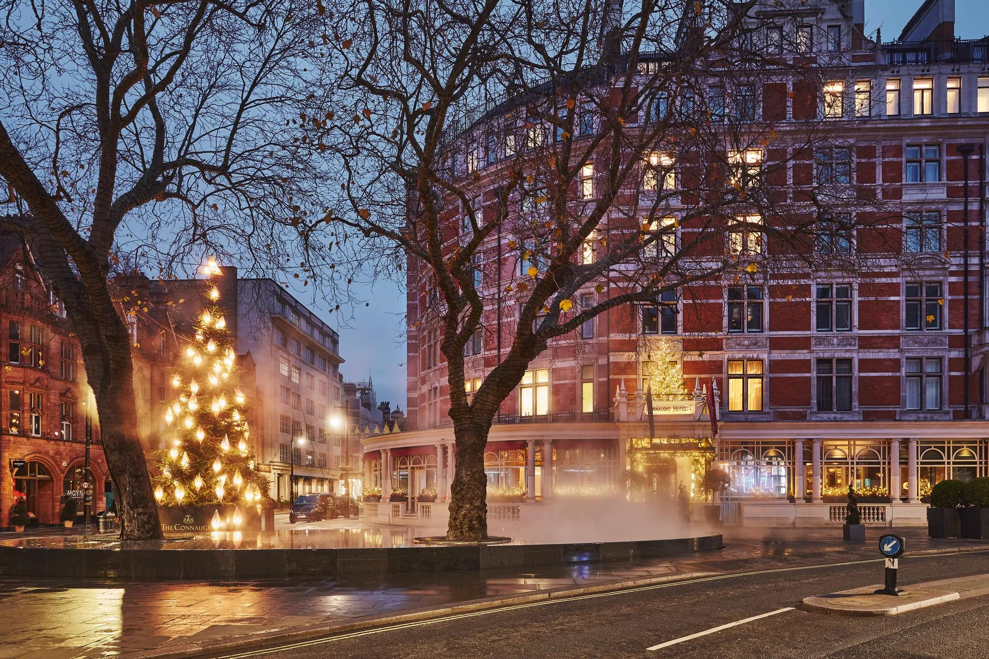 Festive Christmas tree glowing beside a misty fountain outside a grand red-brick hotel at dusk on a wet city street