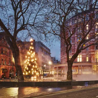 Festive Christmas tree glowing beside a misty fountain outside a grand red-brick hotel at dusk on a wet city street