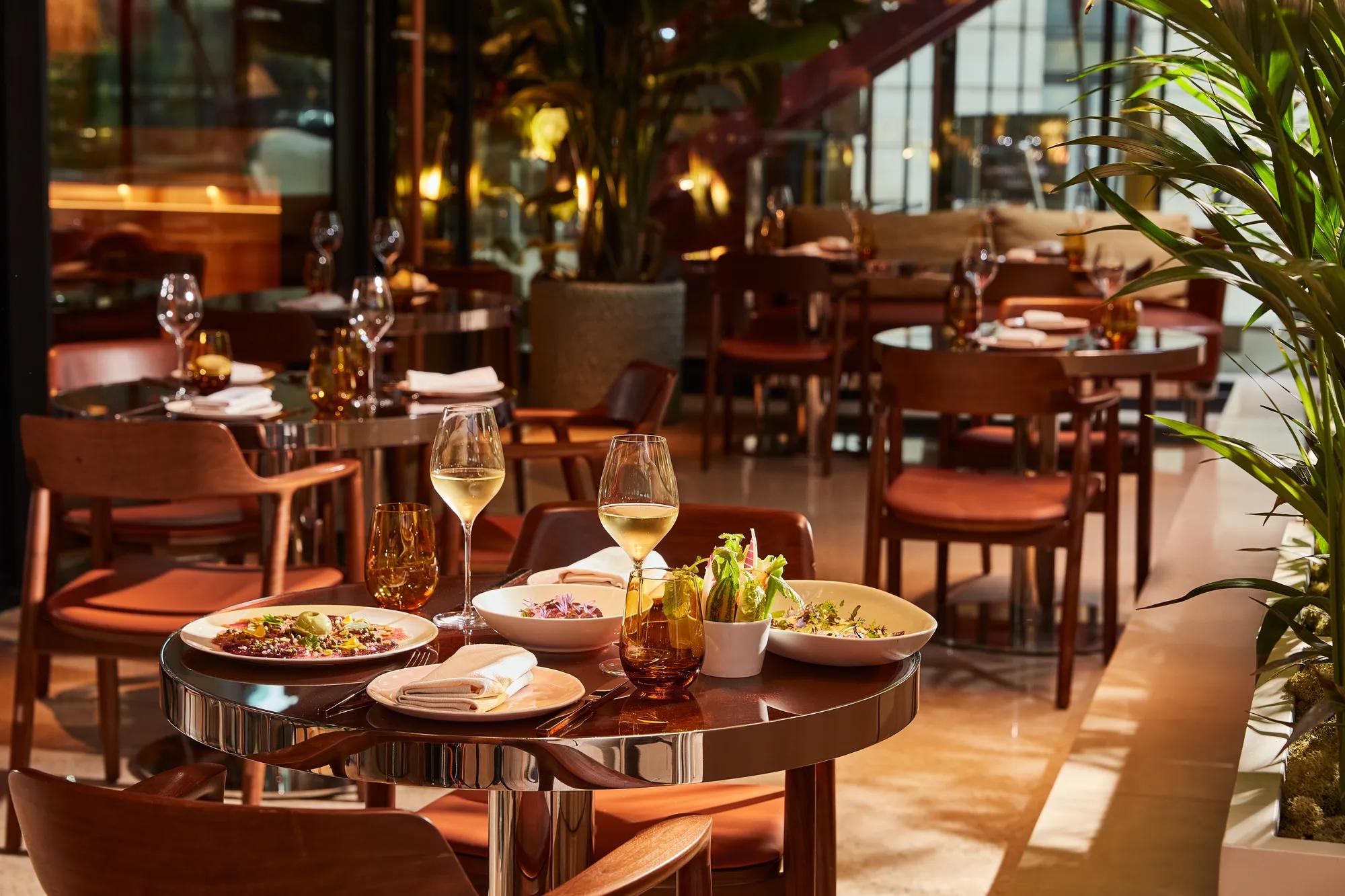Table set for two with white wine, salad, and small plates in a sunlit restaurant with wooden chairs and greenery.