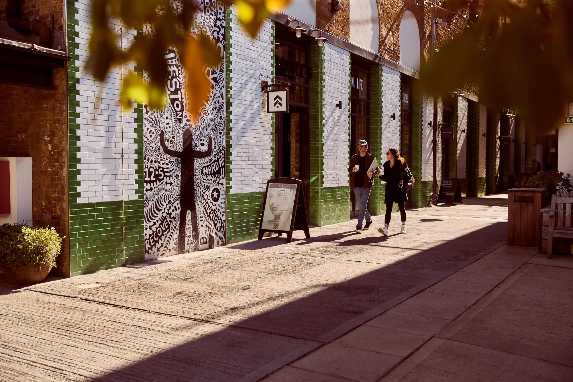 Pedestrians stroll past colourful street art and independent shops beneath railway arches in a sunlit city walkway.