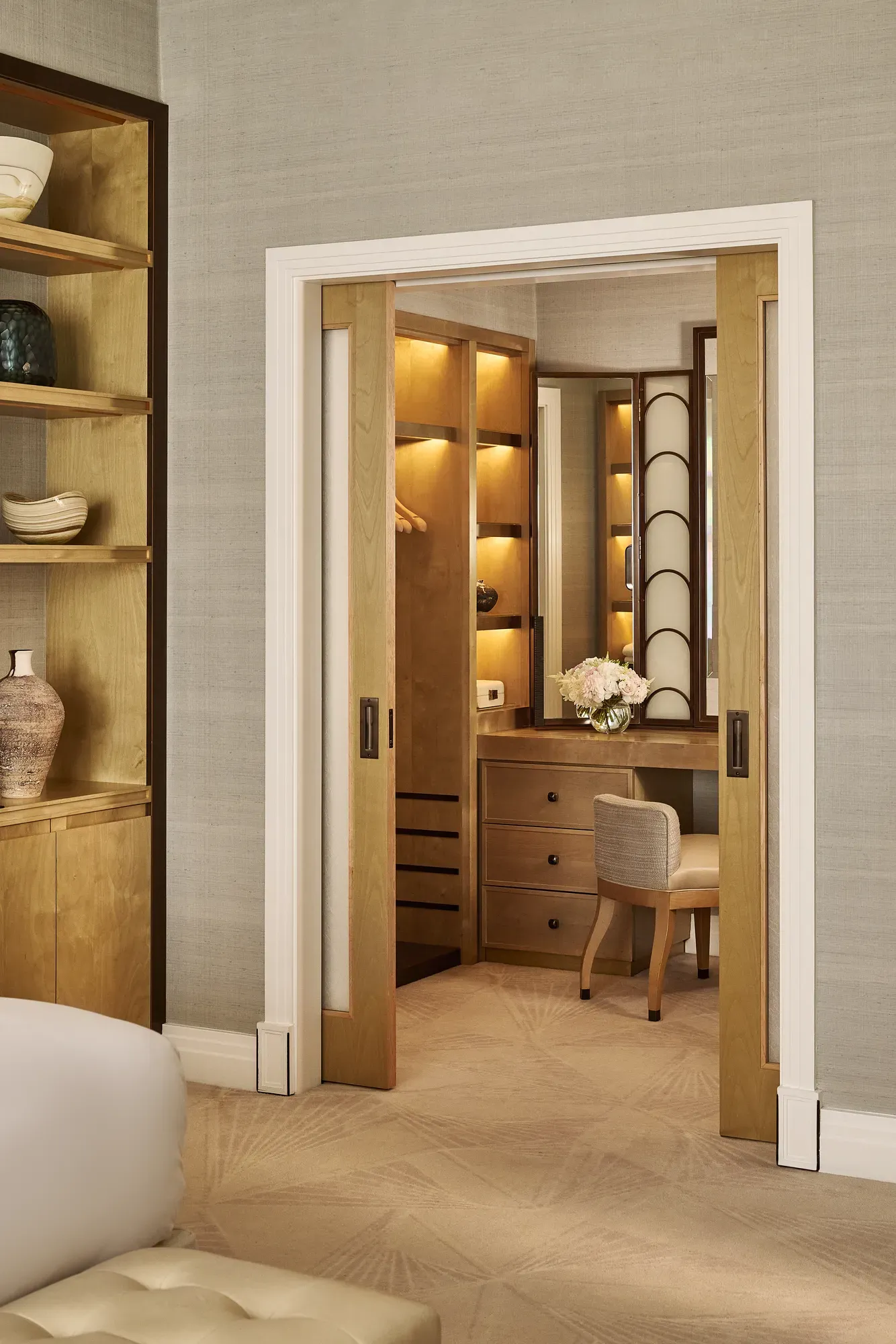 View through sliding doors to walk-in dressing area with illuminated wooden wardrobes, vanity desk, chair, and floral arrangement.