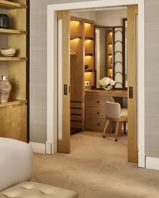 View through sliding doors to walk-in dressing area with illuminated wooden wardrobes, vanity desk, chair, and floral arrangement.