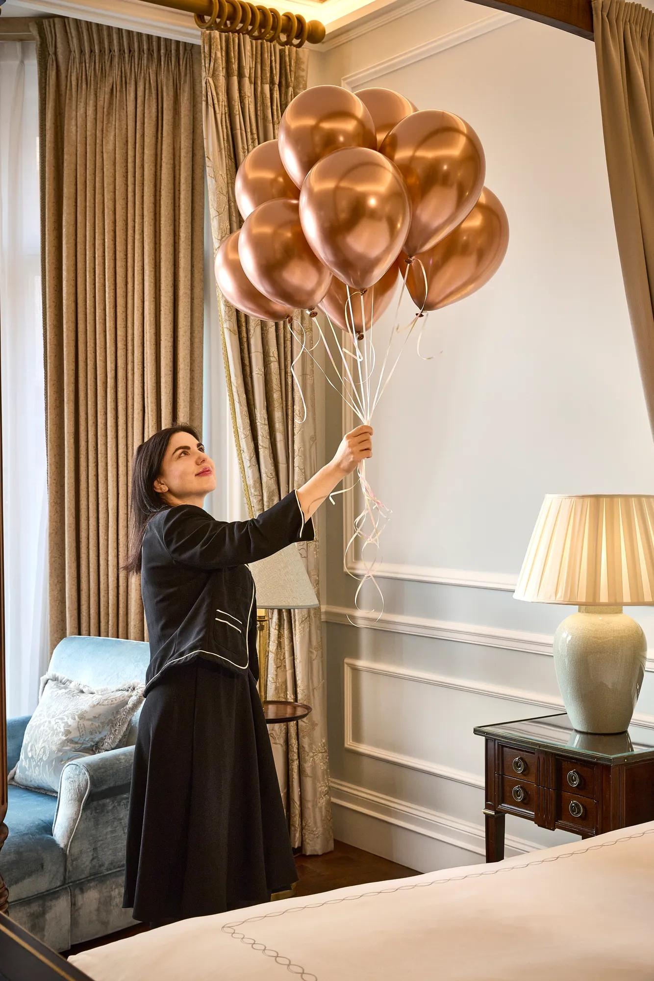 Person in a black outfit holding a cluster of rose-gold balloons in an elegant room with curtains, lamp, and sofa.
