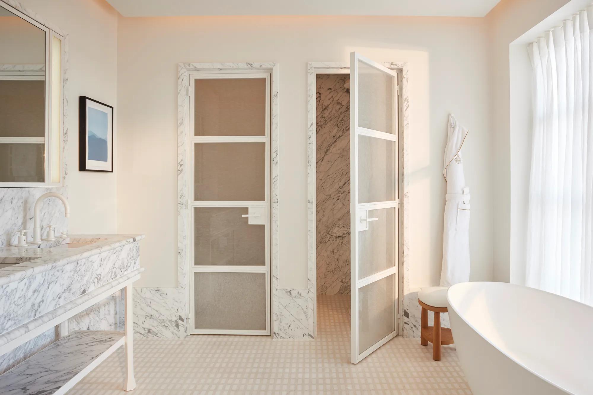 Calm bedroom in soft neutral tones featuring a king-size bed, bedside lamp and flowers, and a view through to a marble bathroom with circular illuminated mirrors.