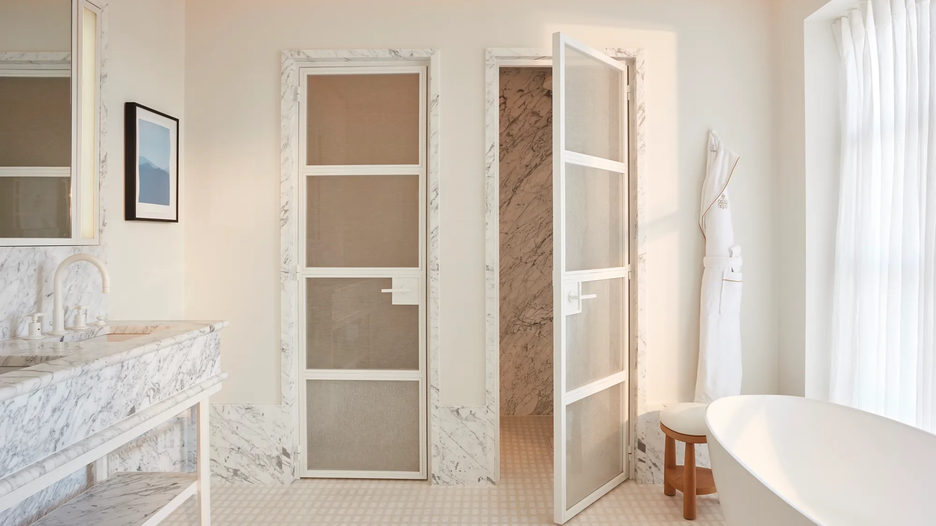 Calm bedroom in soft neutral tones featuring a king-size bed, bedside lamp and flowers, and a view through to a marble bathroom with circular illuminated mirrors.