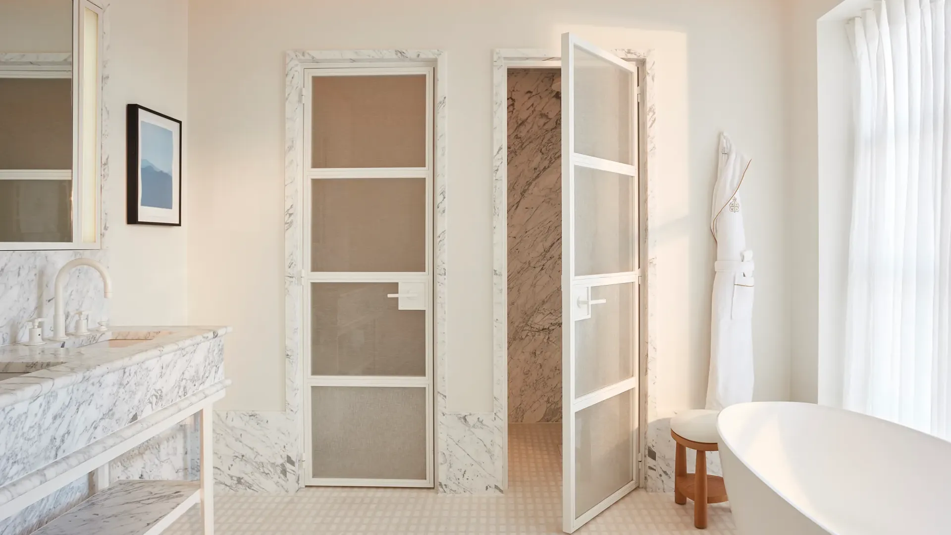 Calm bedroom in soft neutral tones featuring a king-size bed, bedside lamp and flowers, and a view through to a marble bathroom with circular illuminated mirrors.