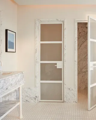 Calm bedroom in soft neutral tones featuring a king-size bed, bedside lamp and flowers, and a view through to a marble bathroom with circular illuminated mirrors.