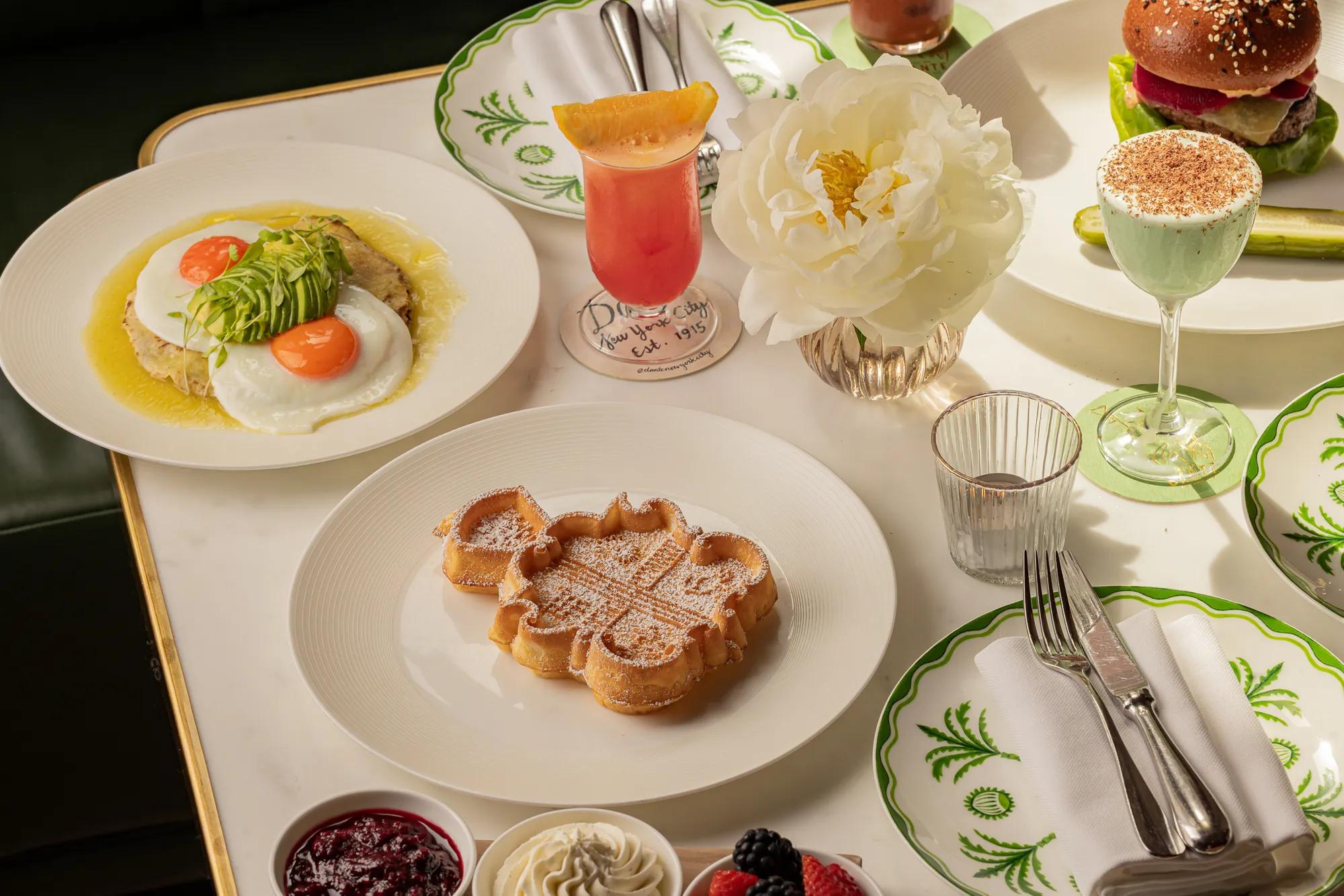 Brunch table with waffle, avocado eggs, burger, cocktails and white flower in a bright restaurant setting.