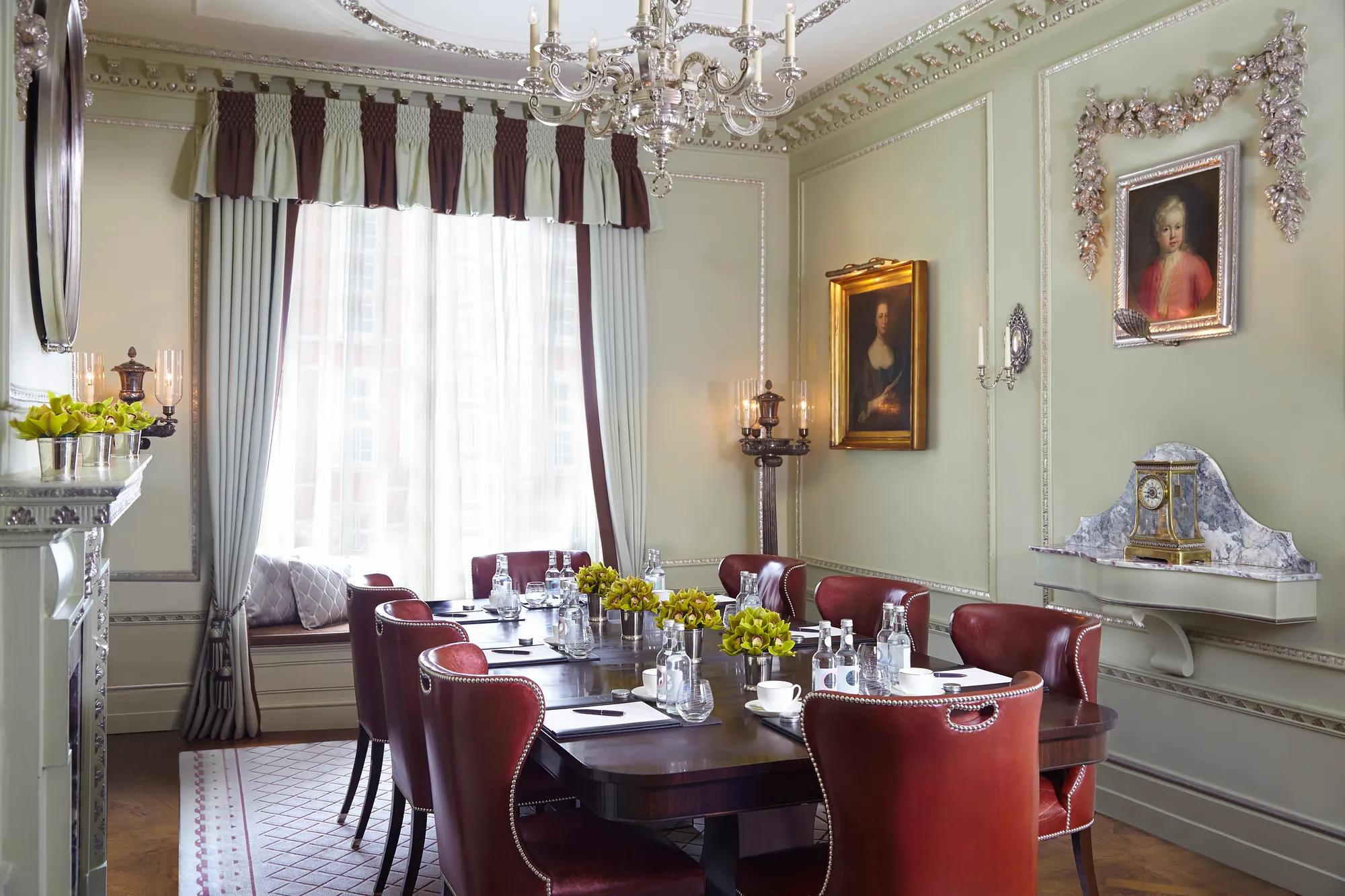Ornate meeting room with a dark wooden table, red leather chairs, notepads and water, framed by chandeliers, classical portraits and soft draped windows.
