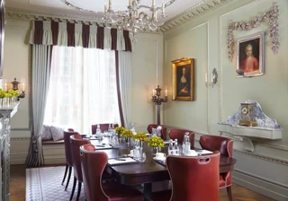 Ornate meeting room with a dark wooden table, red leather chairs, notepads and water, framed by chandeliers, classical portraits and soft draped windows.